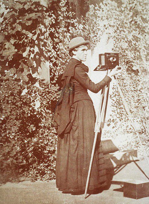 A 19th-century female photographer stands outdoors, dressed in a long black gown and hat, holding a large camera on a tripod amidst lush greenery.