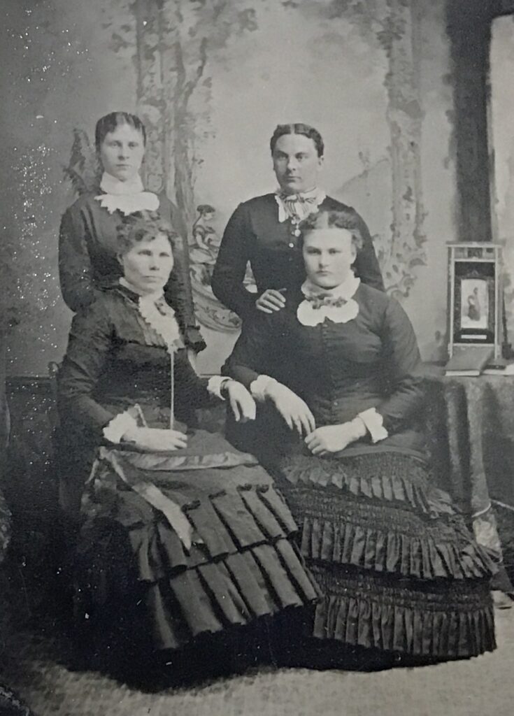 Group portrait of four women in 19th-century Victorian dresses posing in a photography studio with a decorative backdrop.