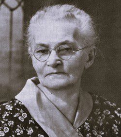 Black-and-white portrait of an elderly woman with short gray hair and glasses, wearing a floral-patterned blouse and a light scarf.