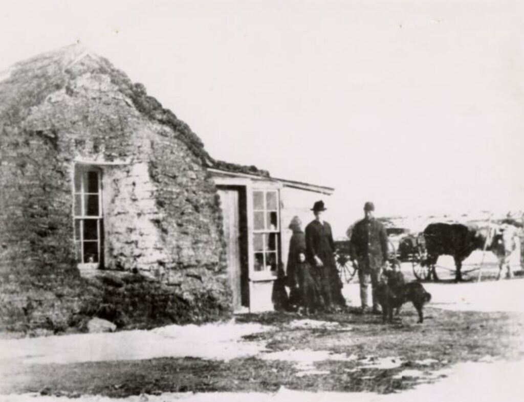 Circular stone hut with a rounded roof and open doorway; a group of people, a dog, and a horse-drawn cart on a bare rural road.