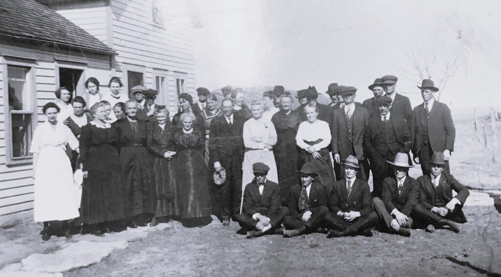 Large group of men and women posing outside a wooden house, in early 1900s clothing (black-and-white photo).
