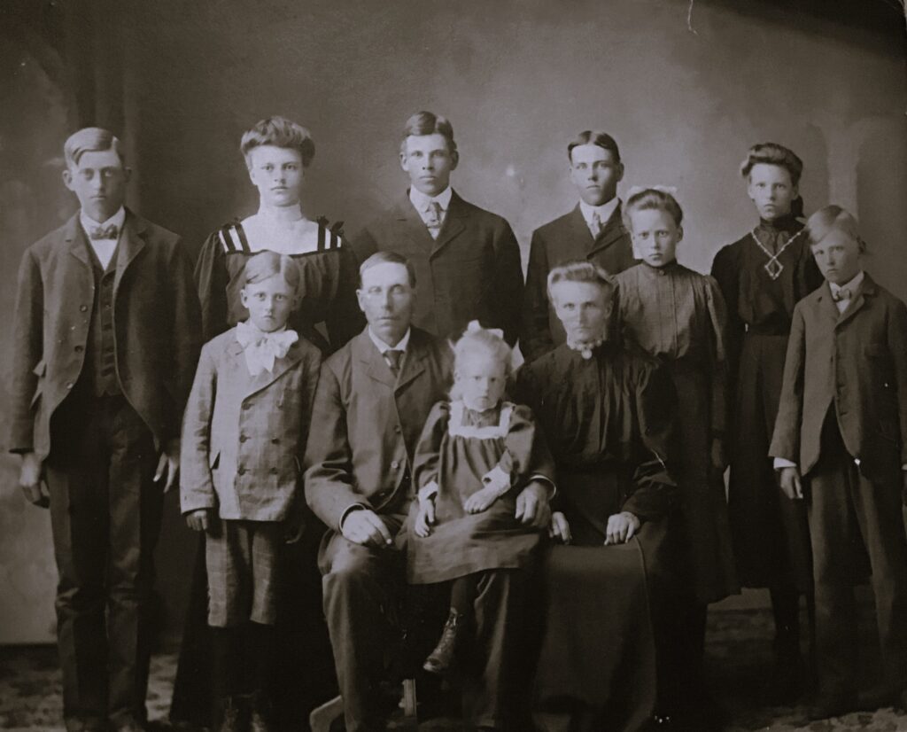 Large black-and-white family portrait: two adults seated center with a young child on the woman's lap, surrounded by several children in formal 19th–early 20th century attire.