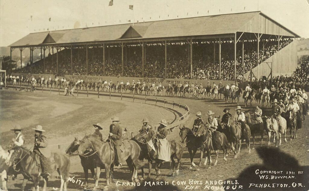 Sepia-toned 1911 photograph of a grand parade at Pendleton Round-Up: riders on horses circle the arena as a packed grandstand overlooks the track; handwritten caption reads 'Grand March Cowboys and Girls – The Round-Up – Pendleton, OR'