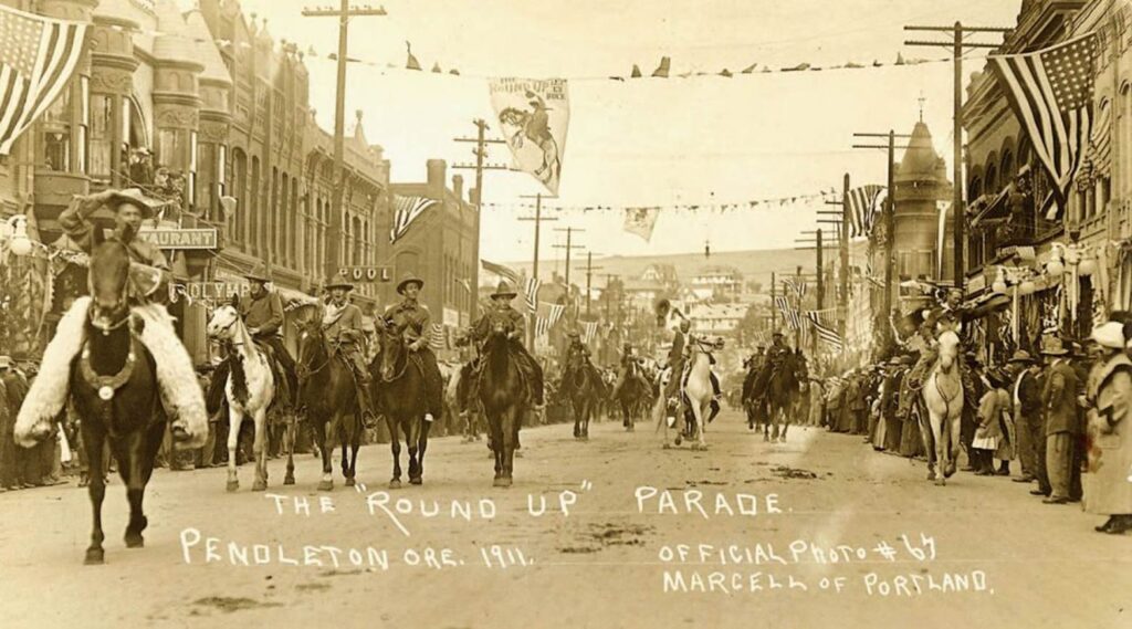 Horse-mounted parade down a dusty main street, Pendleton Round-Up, 1911, with crowds and flags on both sides.